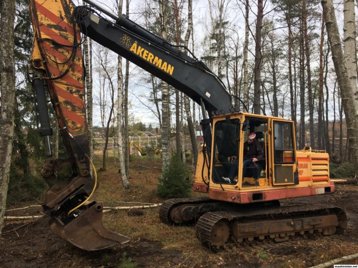 Äldre grävmaskin av märket Åkerman i skogsmiljö med förare i hytten.