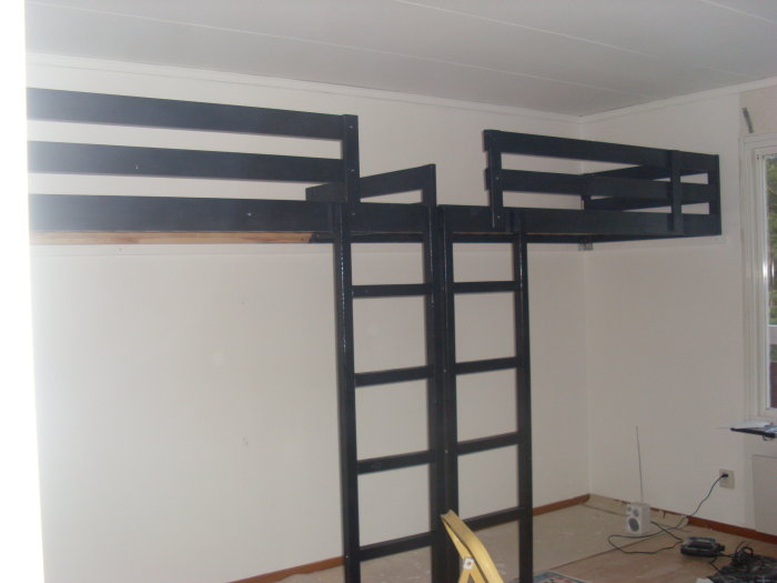 Svart stålkonstruktion av loftbädd med integrerad stege under uppbyggnad i ett rum.