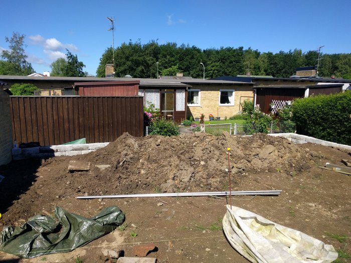 En trädgård under ombyggnad med högar av grävd jord, delvis byggd mur och längder av rör på marken.