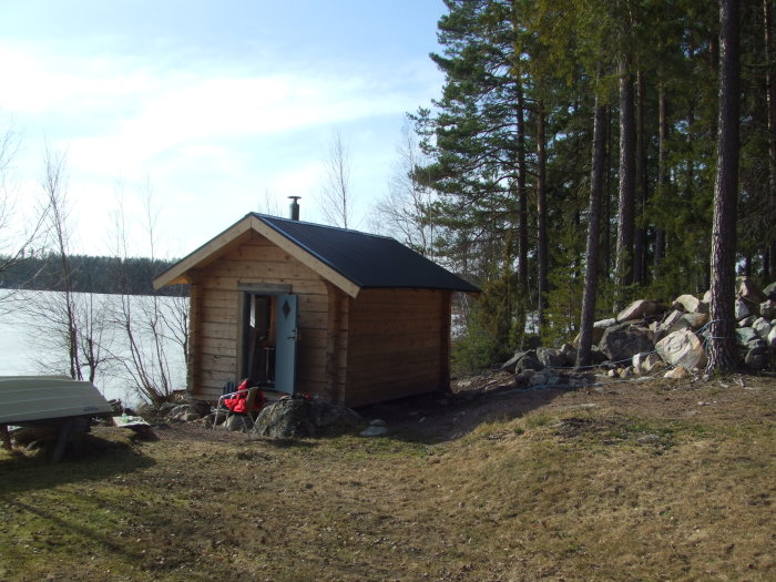 Timmerbastu vid en sjö med öppen dörr, skorsten på taket och omgiven av tallar och klippor.