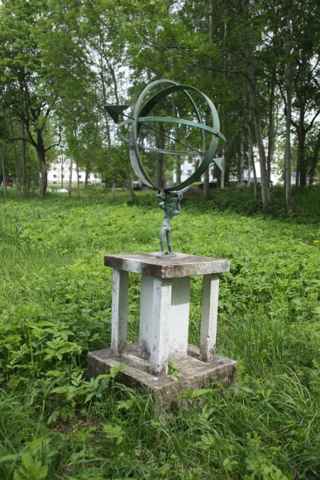 Grönt solur av metall på vit betongfundament i grön omgivning.