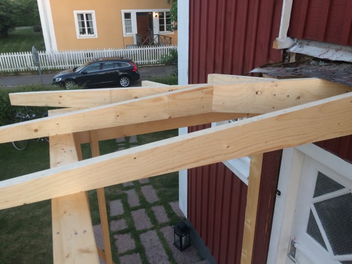 Nykonstruerade träbalkar sammankopplade till en takstom för en veranda, med en röd och gul byggnad i bakgrunden.