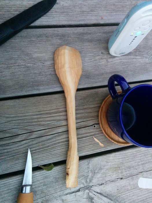 Handsnidad träsked bredvid en morakniv och en blå mugg på ett träbord.