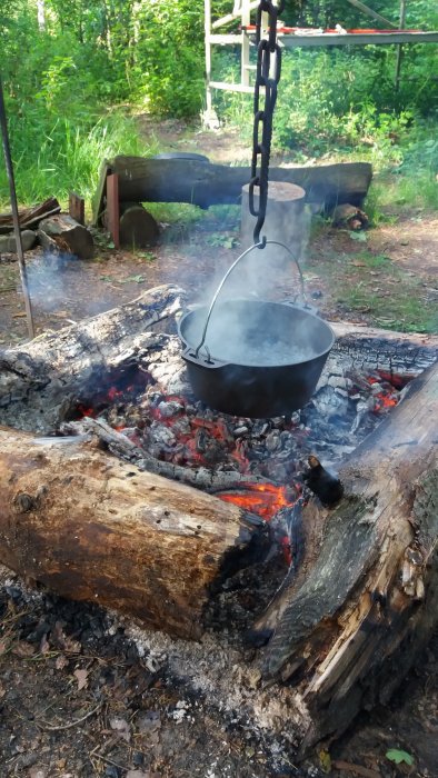 Gjutjärnsgryta hängande över öppen eld mellan två grova trästockar i en skogsglänta.