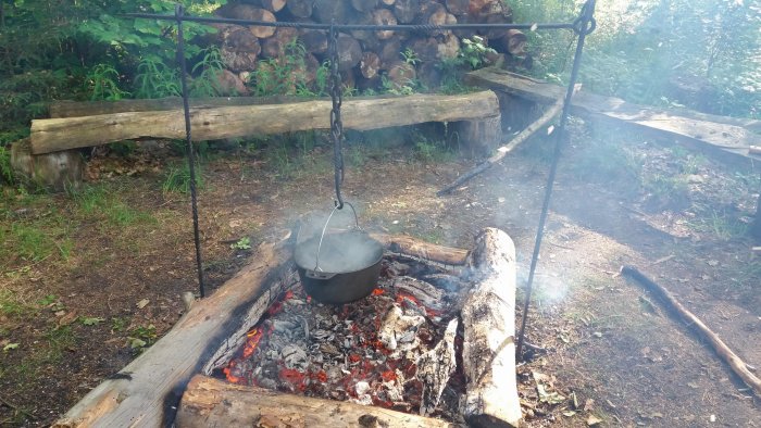 Gjutjärnsgryta som hänger över öppen eld i en lägereldscirkel omgiven av skog.