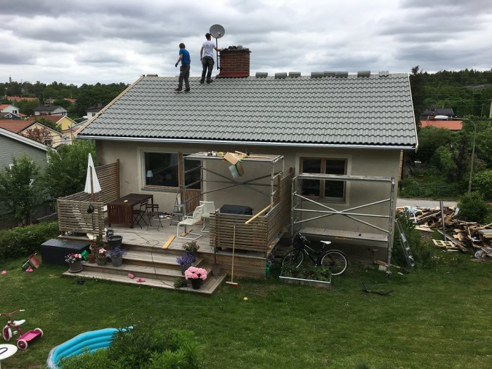 Hus med nytt tak av betongpannor och ställning, två personer på taket, trädgård med leksaker i förgrunden.