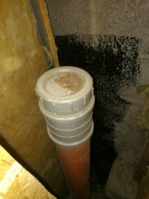 Vertikalt rör med lock, delvis inbyggt bakom isolering och väggskivor i en källare.