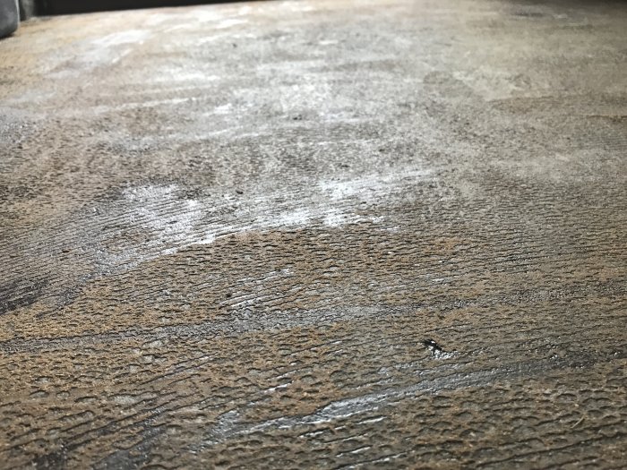 Betonngolv med svarta limrester och bruna mattrester efter borttagning av plastmatta.