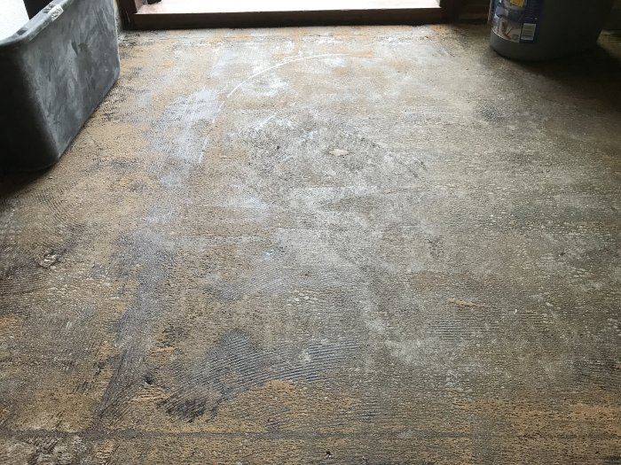 Betonggolv med limrester och brun mattborttagning som förberedelse för klinkerläggning.