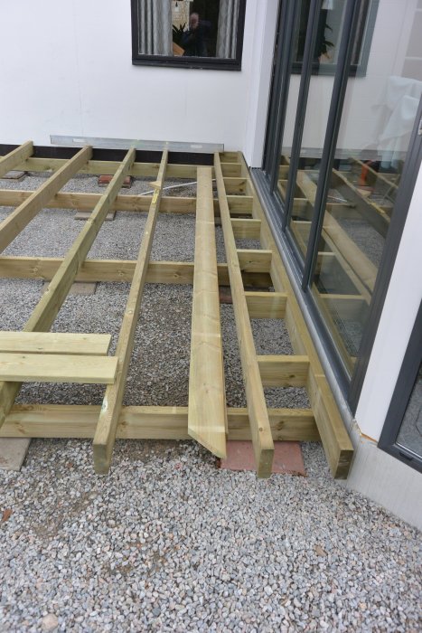 Underlag för trall bredvid glasdörr, träbjälkar och grus, med bredare fris för dränering.