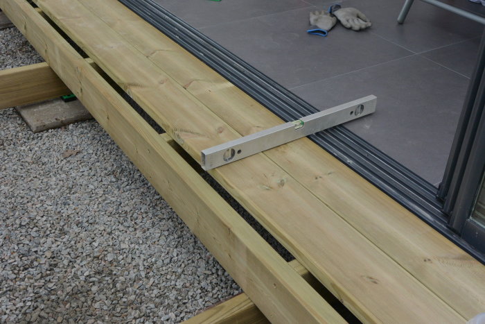 Breda träbrädor vid en skjutdörr med breddat underlag och andvänd trall, inklusive en vattenpass för noggrannhet i byggprojektet.