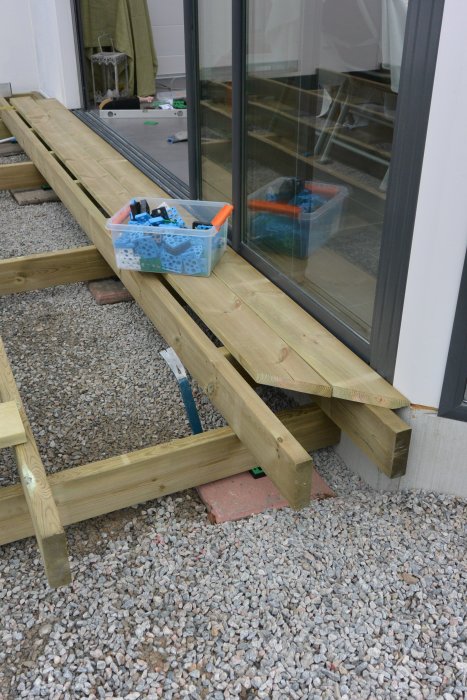 Uppbyggd trätrall framför fönsterdörr med verktygslåda och distansklossar, på singelbredvid gammal cementplatta.