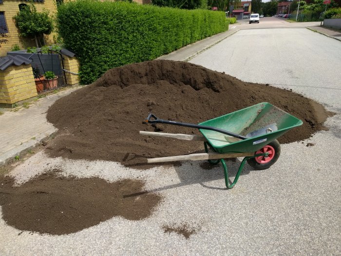 En hög med jord på en uppfart bredvid en grön skottkärra och en spade.