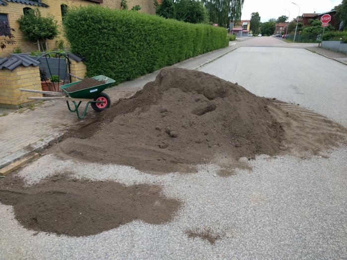 En stor hög med jord levererad på en gata med en tom skottkärra bredvid, redo för ett byggprojekt.