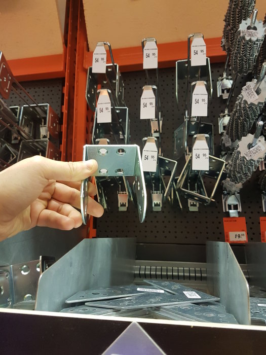 Hand håller en balksko framför ett hyllplan med flera olika balkskor i en bygghandelsbutik.