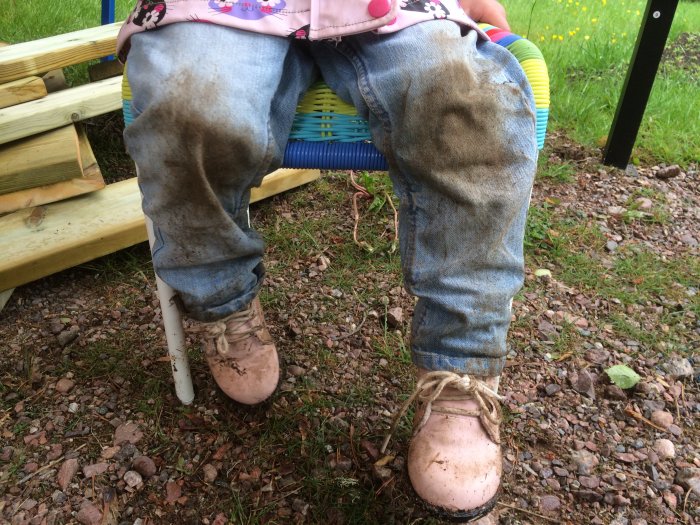 Ett barn sitter på en stol med leriga jeans och rosa stövlar, indikerar utomhuslek och byggprojekt i närheten.