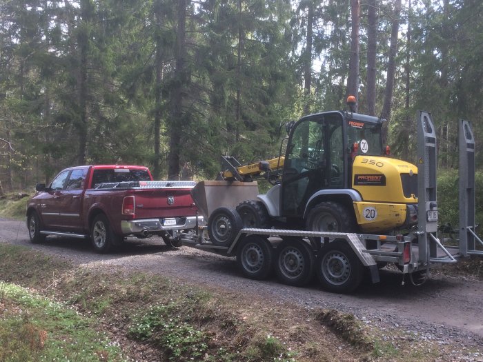 En röd pickup lastar en gul traktor på en släpvagn i skogsmiljö.