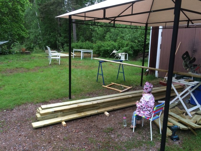 Trädgård med pågående trallbygge, virke på marken, kapmaskin, och barn i regnkläder sitter vid arbetsbänk.