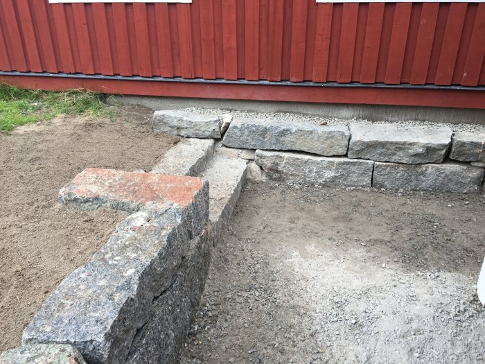 Stenbelagd trädgårdsväg under konstruktion intill en röd trävägg med ett steg av stenblock.