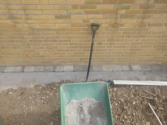 Hörn av en nybyggd stenmur med en spade lutad mot den och en skottkärra med grus i förgrunden.