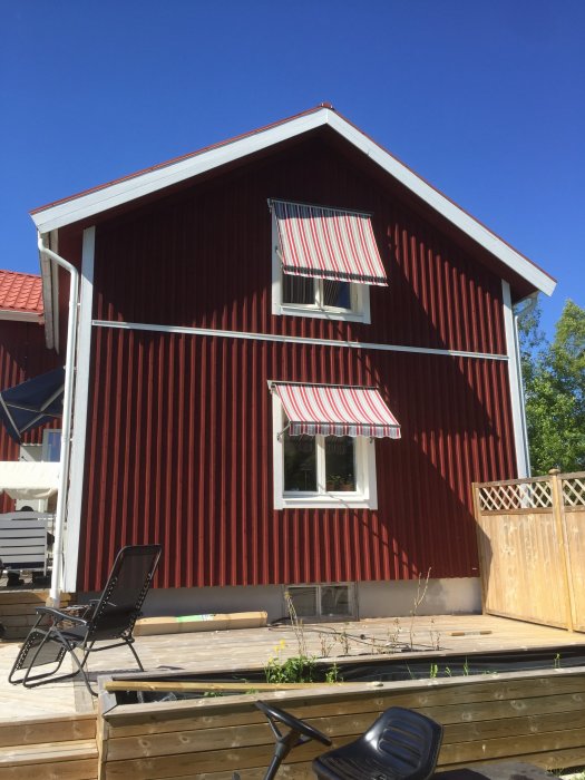 Röd trävilla med två randiga markiser, en på varje våningsplan, under en klarblå himmel.