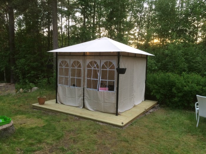Vit paviljongtält med sidor och fönster monterad på en träplattform ute i skogstomt vid skymning.