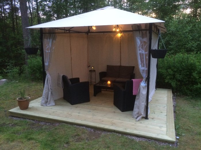 Utomhus vardagsrum med soffgrupp under paviljong på trädäck omgiven av grönska i skymning.