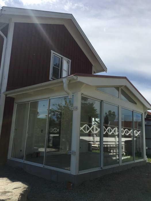 Nästan färdigt uterum med glasade sektioner intill rött trähus, saknar stuprör och inre elarbeten.