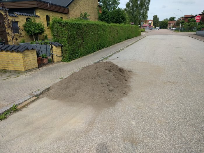 Hög med jord på en asfalterad väg vid kantsten nära en häck och ett gult tegelhus.