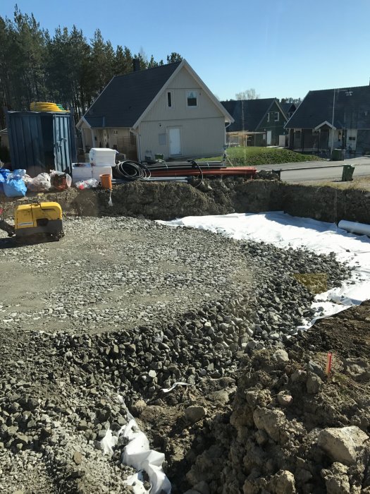 Stor grushög i en grop vid ett byggområde med hus och byggmaterial i bakgrunden.