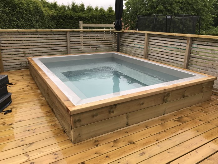 Trädäck med inbyggd ovanmarkspool, trästaket i bakgrunden, våta ytor som antyder regn.