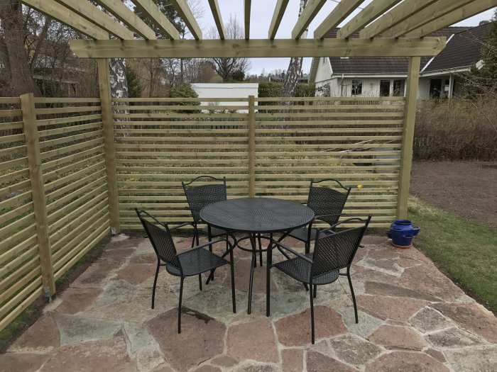 Nybyggd uteplats med en pergola, en rund bord- och stolgrupp på stenplattor, redo för sommarkvällar.