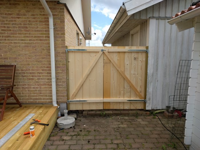 Nybyggd trägrind mellan hus och skjul med verktyg och material på marken.