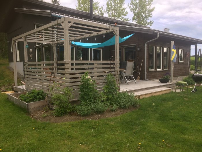 En trätrall vid ett hus med pergola, solskydd, trädgårdsland och en svensk flagga.