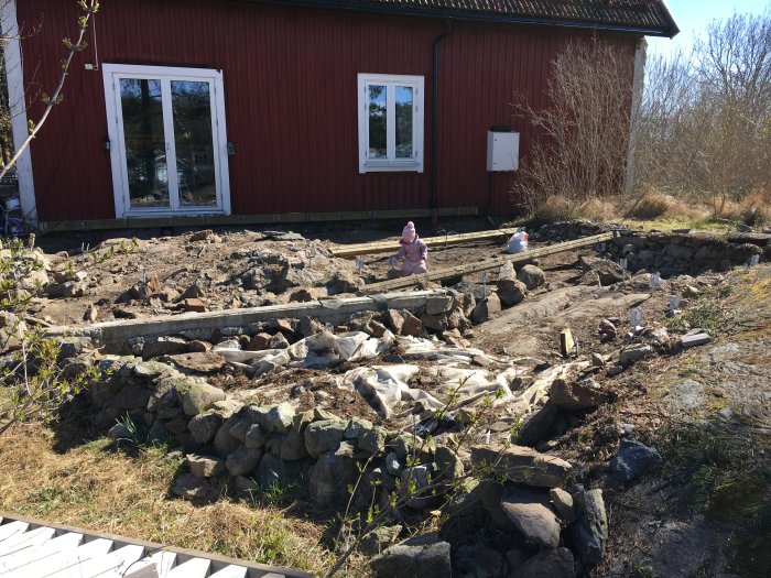 Markarbete med synlig sten och grus framför rött hus, trästaket och två små barn.