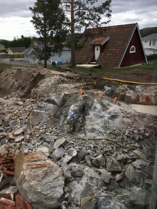 Markarbete med synliga simplex sprängkapslar i berg och sprängsten framför röd stuga.