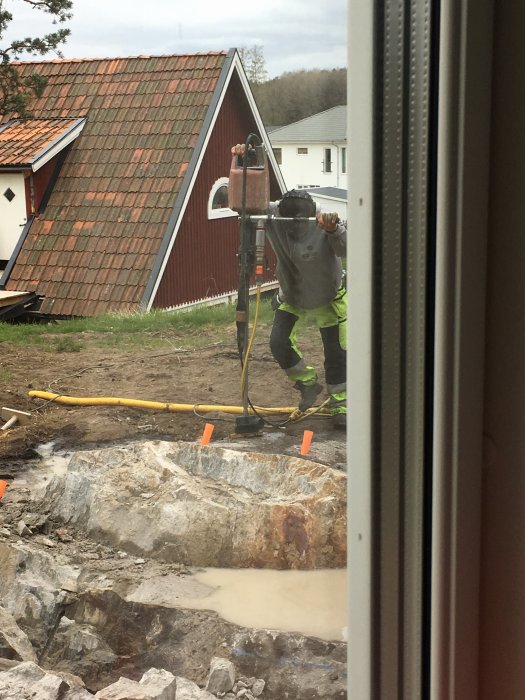 Arbetare vid markarbete använder simplex för att spräcka sten, hus i bakgrunden.