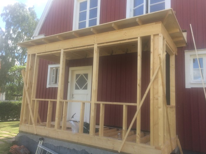 Nybyggd träkonstruktion för veranda med plywood-sarg på röd husfasad under blå himmel.