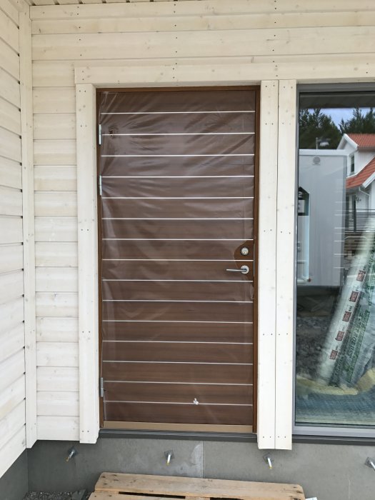 Nyinstallerad brun ytterdörr på ett hus under konstruktion, skyddad med plast, omgiven av vit panel.