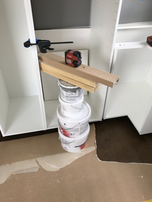 Improviserat laserstativ gjort av träplanka och färgburkar i ett halvfärdigt kök under renovering.