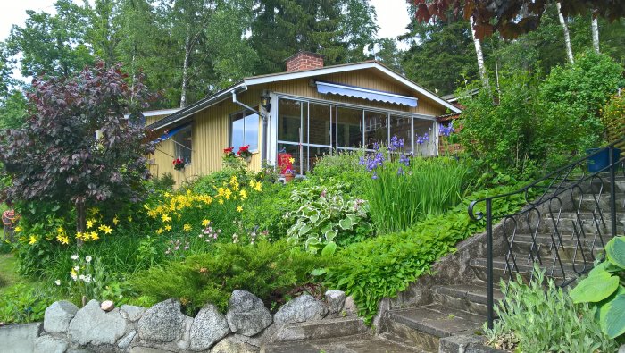 En mångfärgad trädgård med blommor vid ett gult hus med veranda och stentrappa.