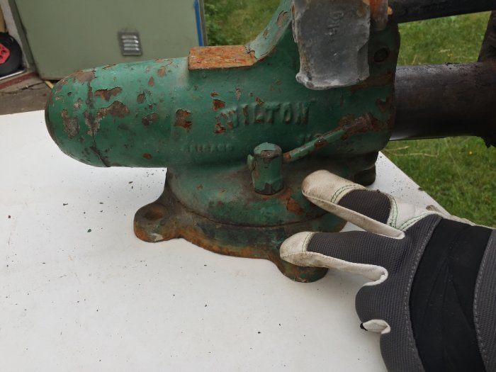 Hand i skyddshandske håller i gammalt grönt skruvstäd med rost och avflagad färg.
