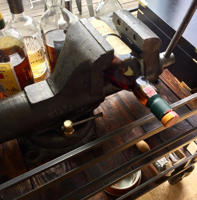 Restaurerat gammalt Wilton-skruvstäd med mässingsdetaljer, flankerat av whiskyflaskor på trähylla.