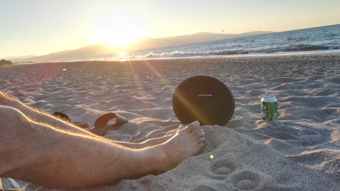 Harman/Kardon högtalare på stranden vid solnedgång med sandiga fötter och en ölburk.