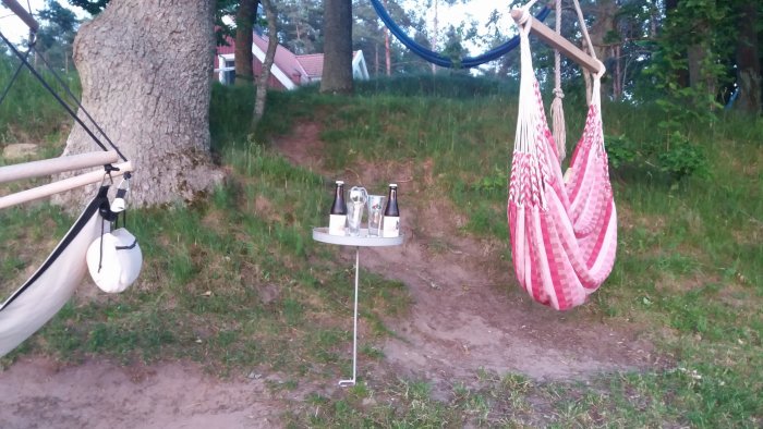 Hängmatta och rosa hängstol med ett sidobord med ölflaskor och glas i trädgården vid solnedgång.