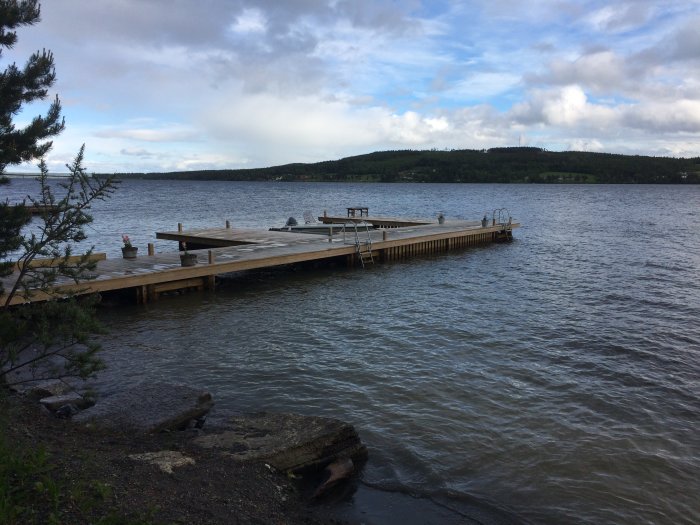 Brygga intill en sjö med högre vattennivå omgiven av grönska och molnig himmel.