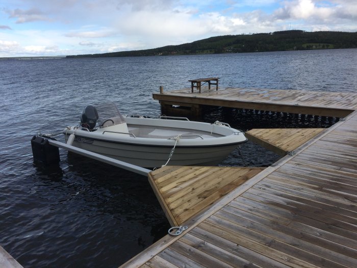 Motorbåt förtöjd vid en träbrygga på en sjö med stigande vattennivå och en bänk på kajen.