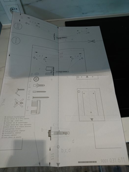 Monteringsinstruktion för köksfläkt med tydliga diagram och delbeteckningar, ligger på ett bord.