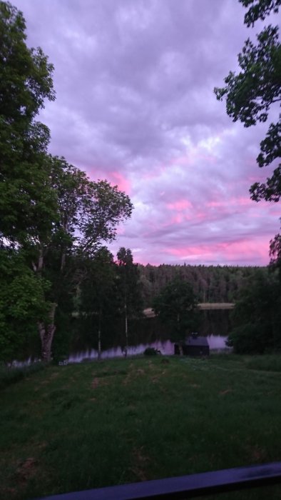 Utsikt från ett sovrumsfönster mot söder med träd, gräsmatta och rosa himmel över en sjö.