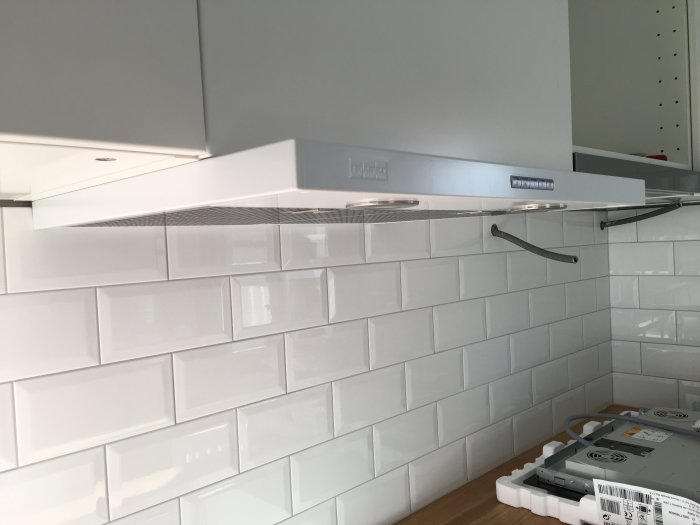 Nymonterad vit köksfläkt ovanför vit kakelvägg med spisunderdel synlig på bänk.
